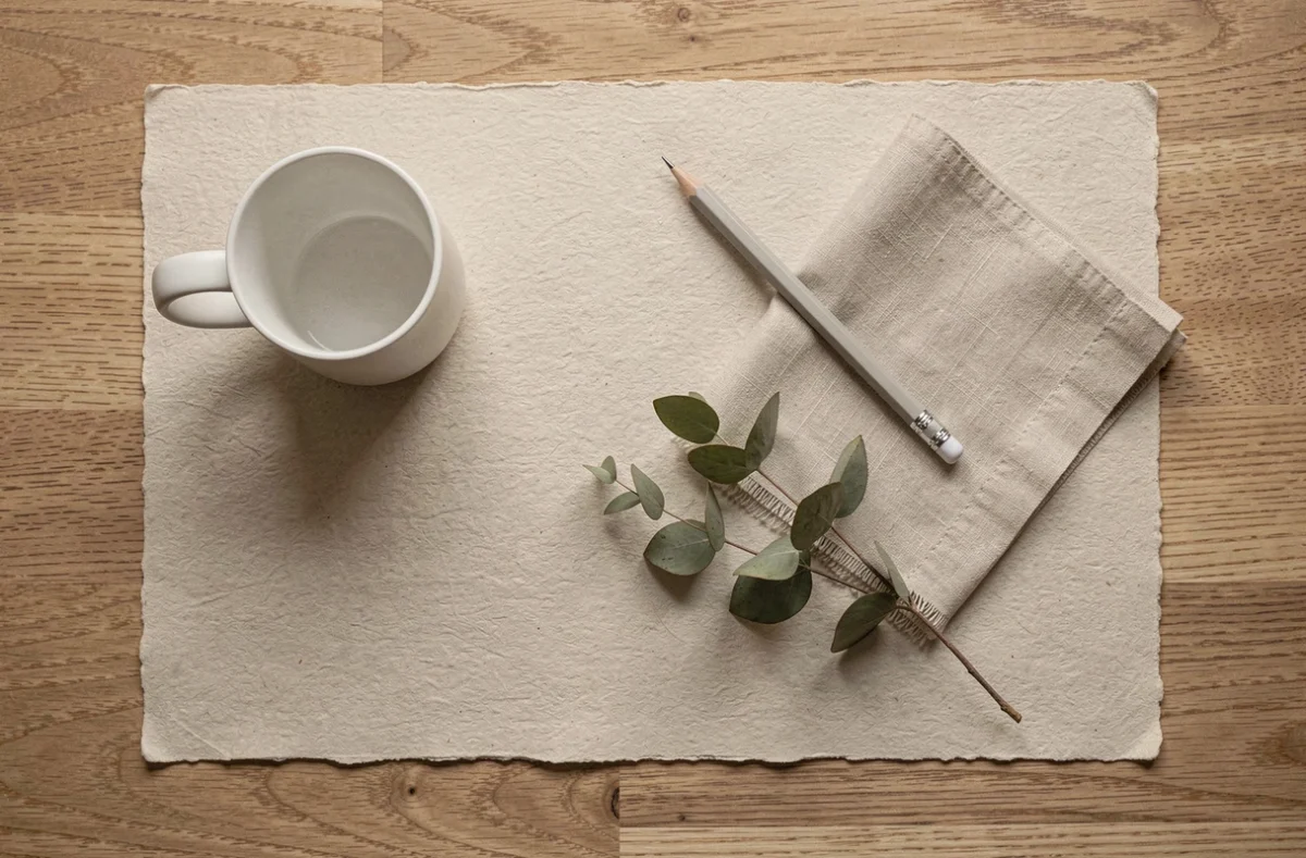 Flat lay minimaliste sur papier beige texturé avec mug blanc mat, crayon et tissu lin écru sur bureau en chêne naturel